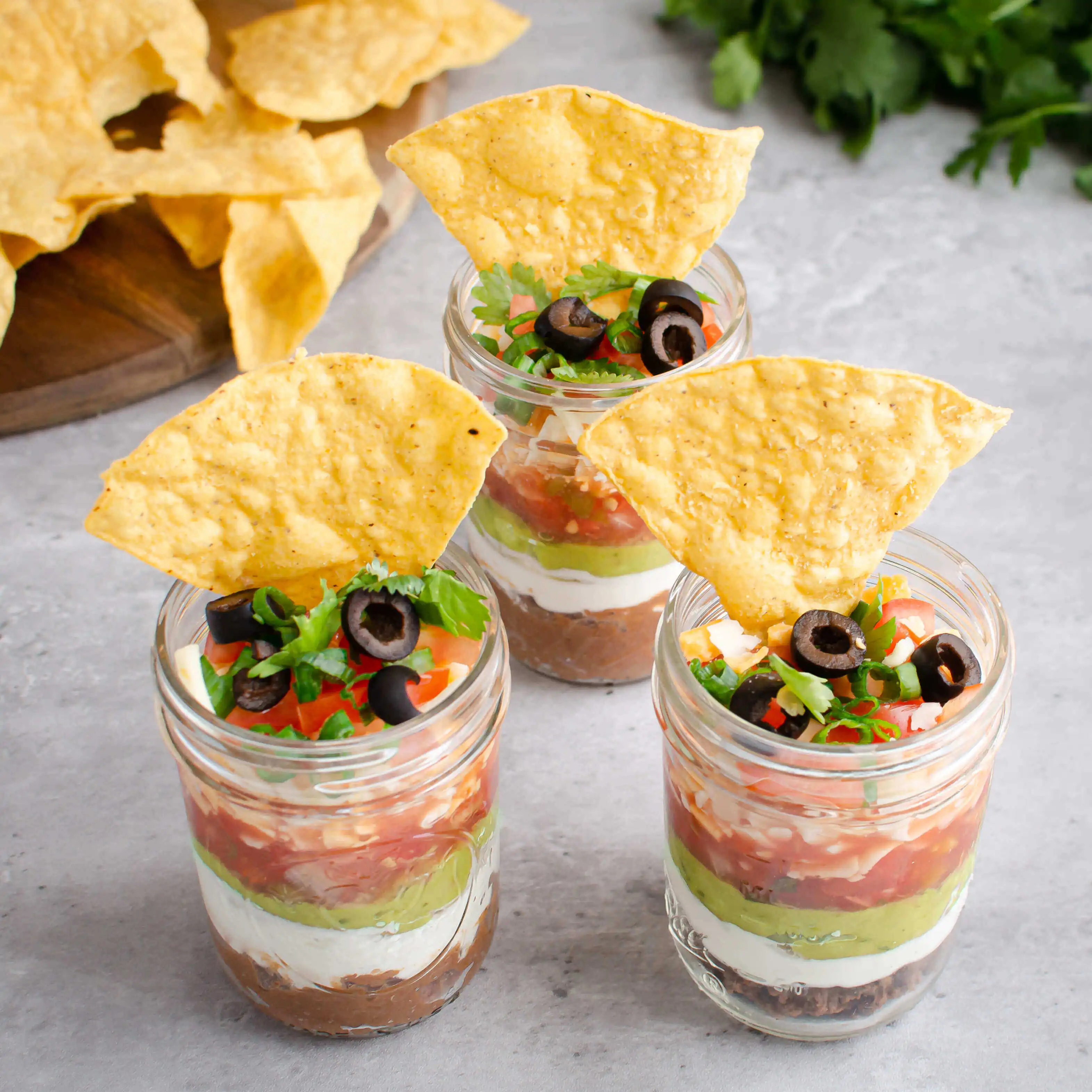 Three 7 Layer Taco Dip cups with tortilla chips.